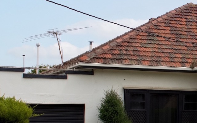 A close crop of a tiled roof, a TV aerial and a power line against a pale blue sky with soft cloud, with clean edges